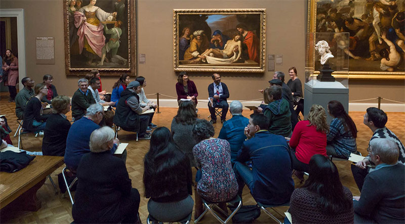 nikhil and Nenette seated with a large circle of people in a gallery. Three large oil paintings hang on the wall behind them.