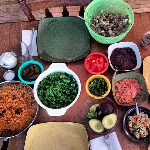 A birdseye view photo of a table of food. There's a bowl of rice, a bowl of cilantro, tomatoes, black beans, halved avocados, cooked pork and chicken, salt, and empty plates and glasses.